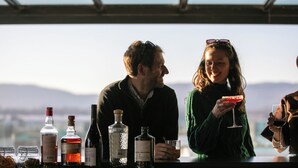 A bar area with various bottles and glasses, a person holding a glass, and a scenic view in the background.