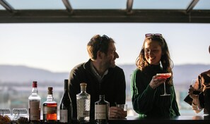 Uma área de bar com diversos copos e garrafas, uma pessoa segurando um copo, e uma vista panorâmica ao fundo.