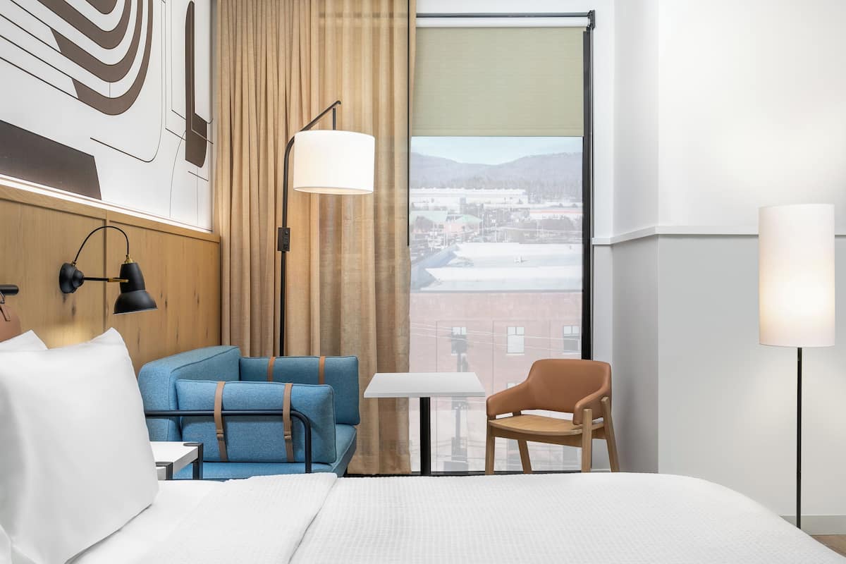 A hotel room with a bed, a blue sofa, a wooden chair, a small table, and a large window with a view of buildings and mountains.