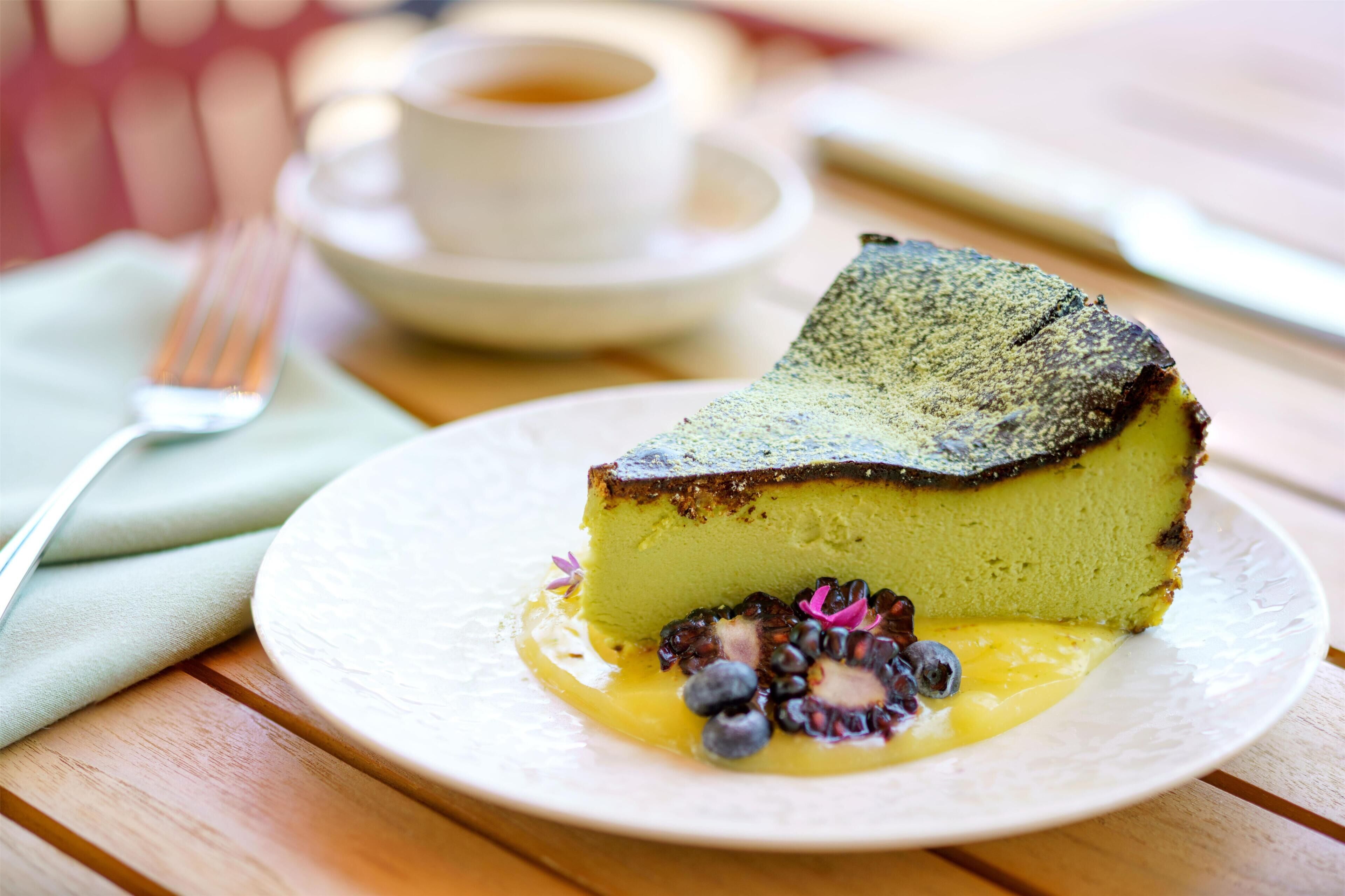 A slice of cheesecake on a white plate with blueberries and a dollop of sauce, accompanied by a cup of coffee on a saucer.