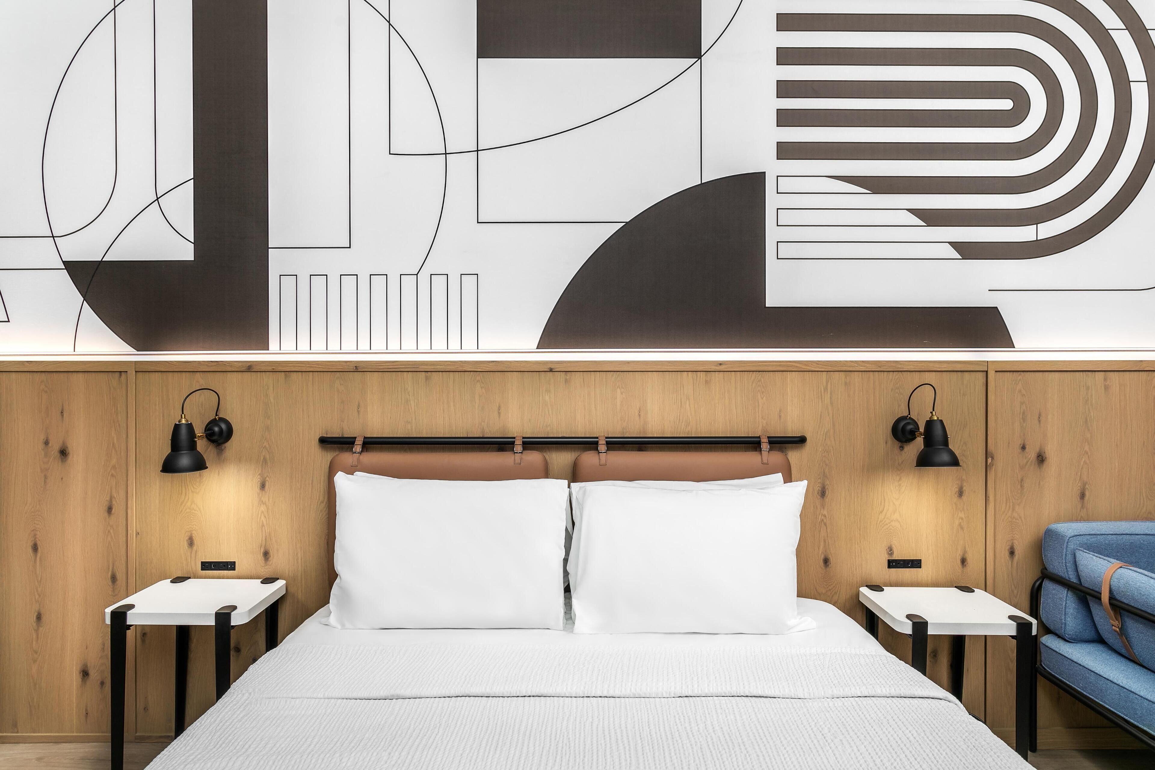 Une chambre d’hôtel moderne avec une tête de lit en bois, une grande œuvre d’art abstraite accrochée au mur, deux tables de chevet avec des lampes, un lit avec du linge de lit blanc et un fauteuil bleu recouvert de tissu.