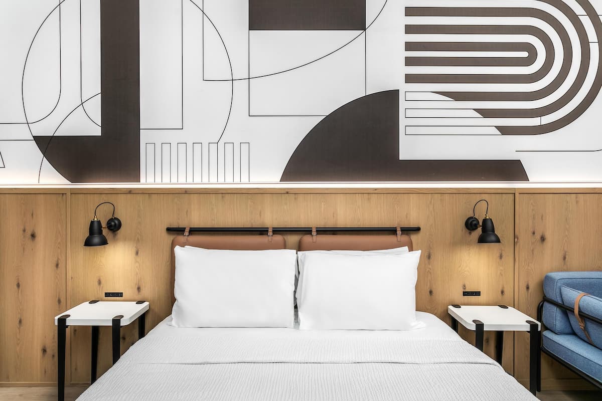 A modern hotel room with a wooden headboard, a large abstract wall art, two bedside tables with lamps, a bed with white linens, and a blue upholstered chair.