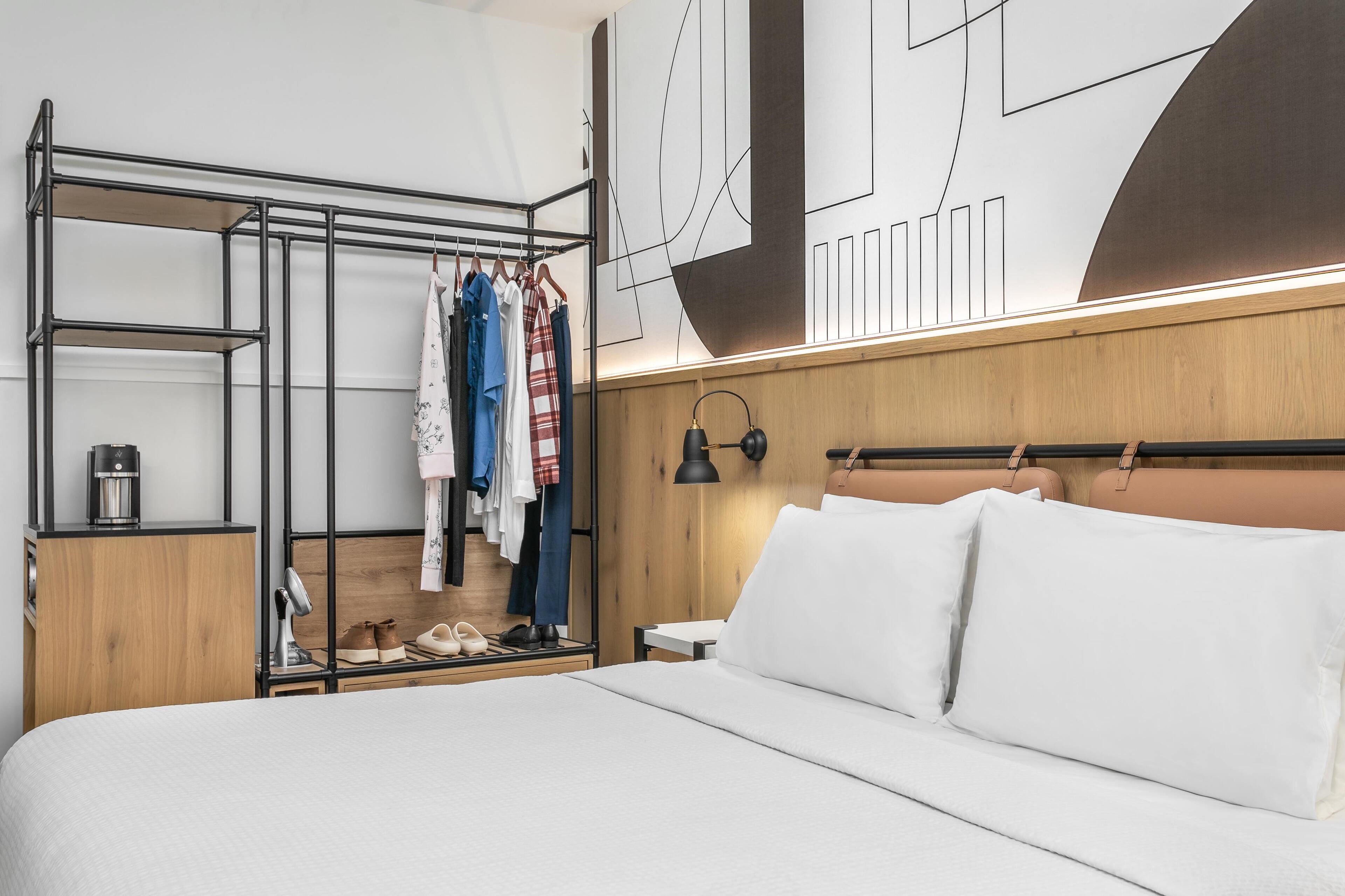 Ett modernt hotellrum med en säng, en garderob i trä, en spegel och en vägg prydd med abstrakt konst.