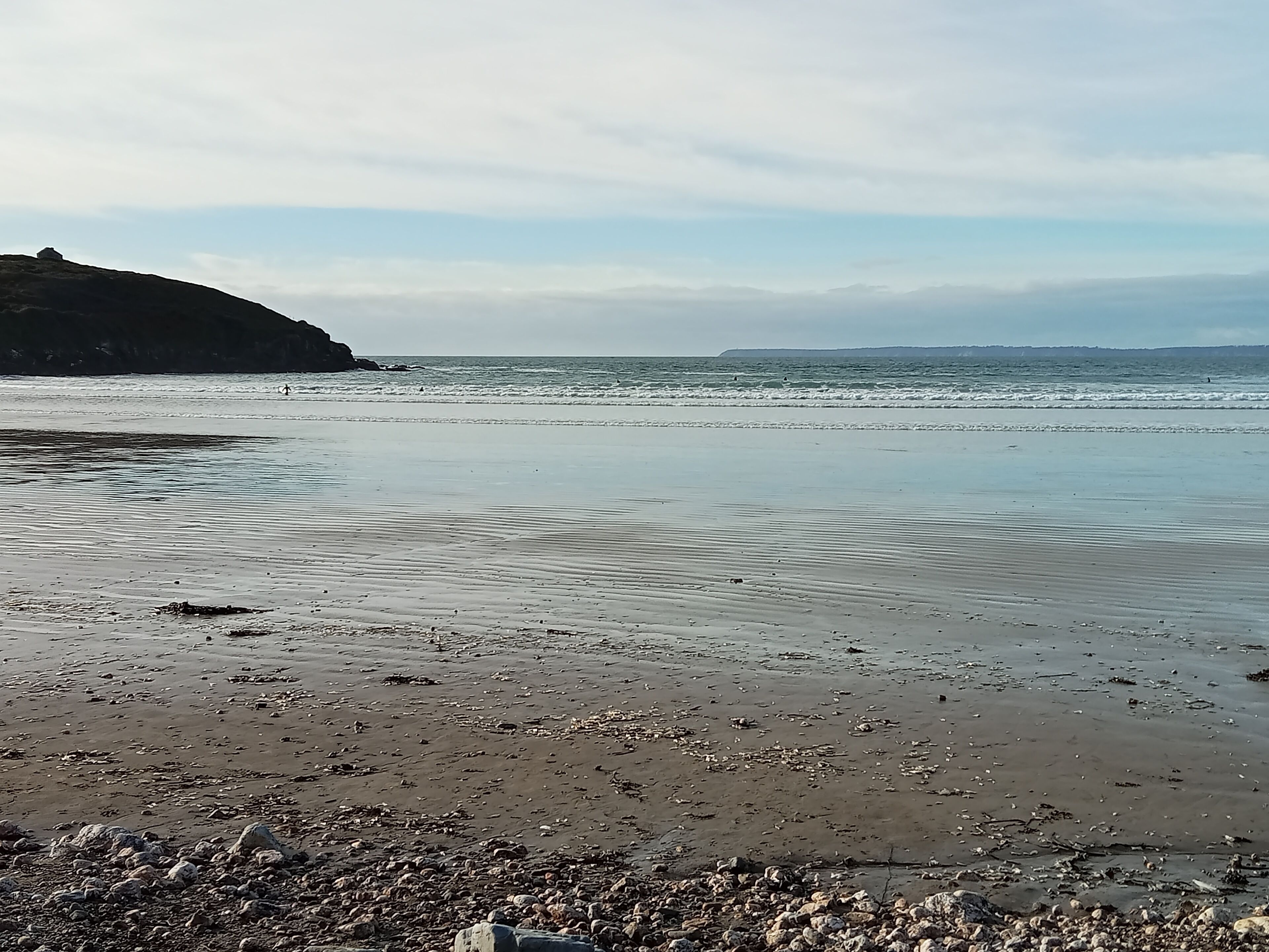 Une plage de galets et de rochers, avec une eau calme et une côte rocheuse qui s’étend au loin.
