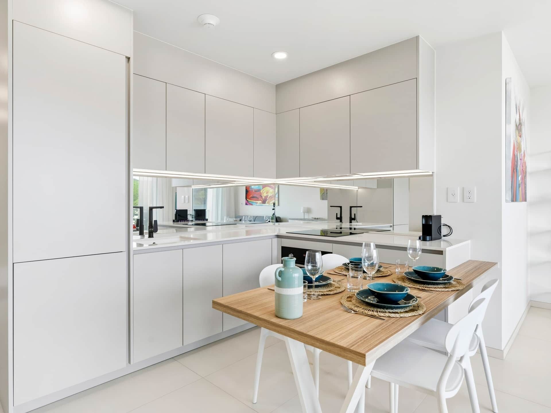 Una cocina moderna con armarios blancos, una mesa de comedor de madera y un espejo grande sobre la encimera.