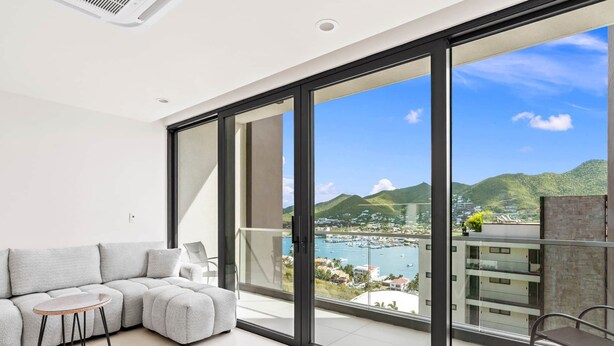 A modern living room with a grey sectional sofa, a small round table, and a balcony with a view of a marina and hills. - D416 – Relaxing apartment with Balcony (Sint Maarten)