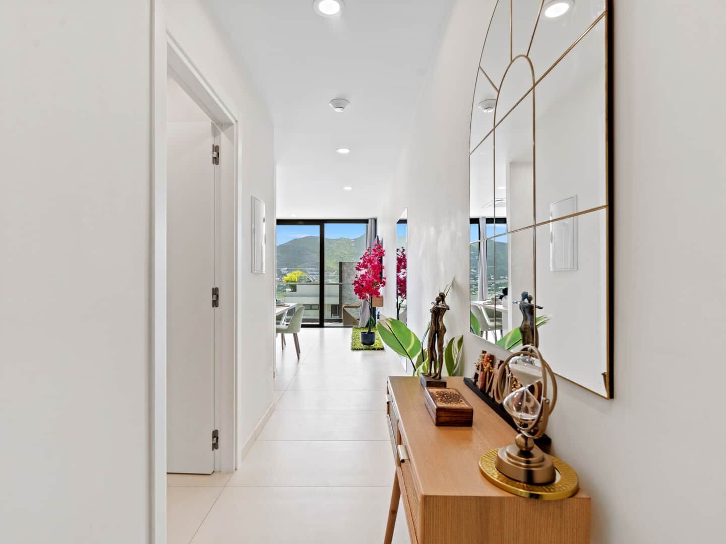 A modern hallway with a wooden console table, a large mirror, and a view of a balcony with plants and a mountain in the distance.