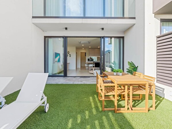 Un patio exterior moderno con una mesa y sillas de madera, una silla lounge blanca y una puerta de vidrio que da a una cocina y sala de estar.