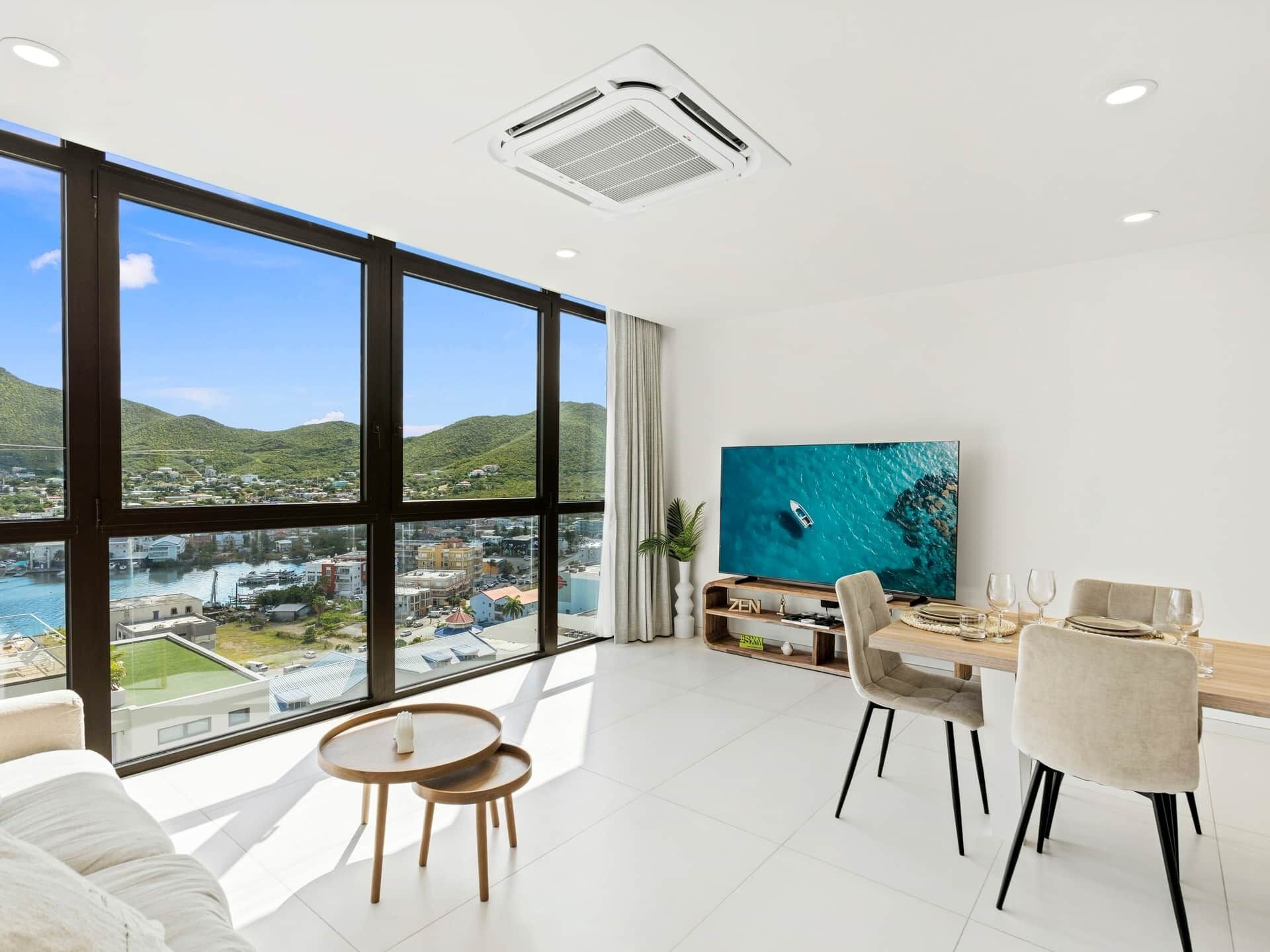A modern living room with a dining area, large windows, and a view of a coastal town.