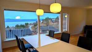 Ein Essbereich mit Tisch und Stühlen, große Fenster mit Blick auf einen See und Gebäude, sowie hängende Pendelleuchten.