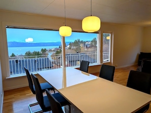 A dining area with a table and chairs, large windows with a view of a lake and buildings, and hanging pendant lights.