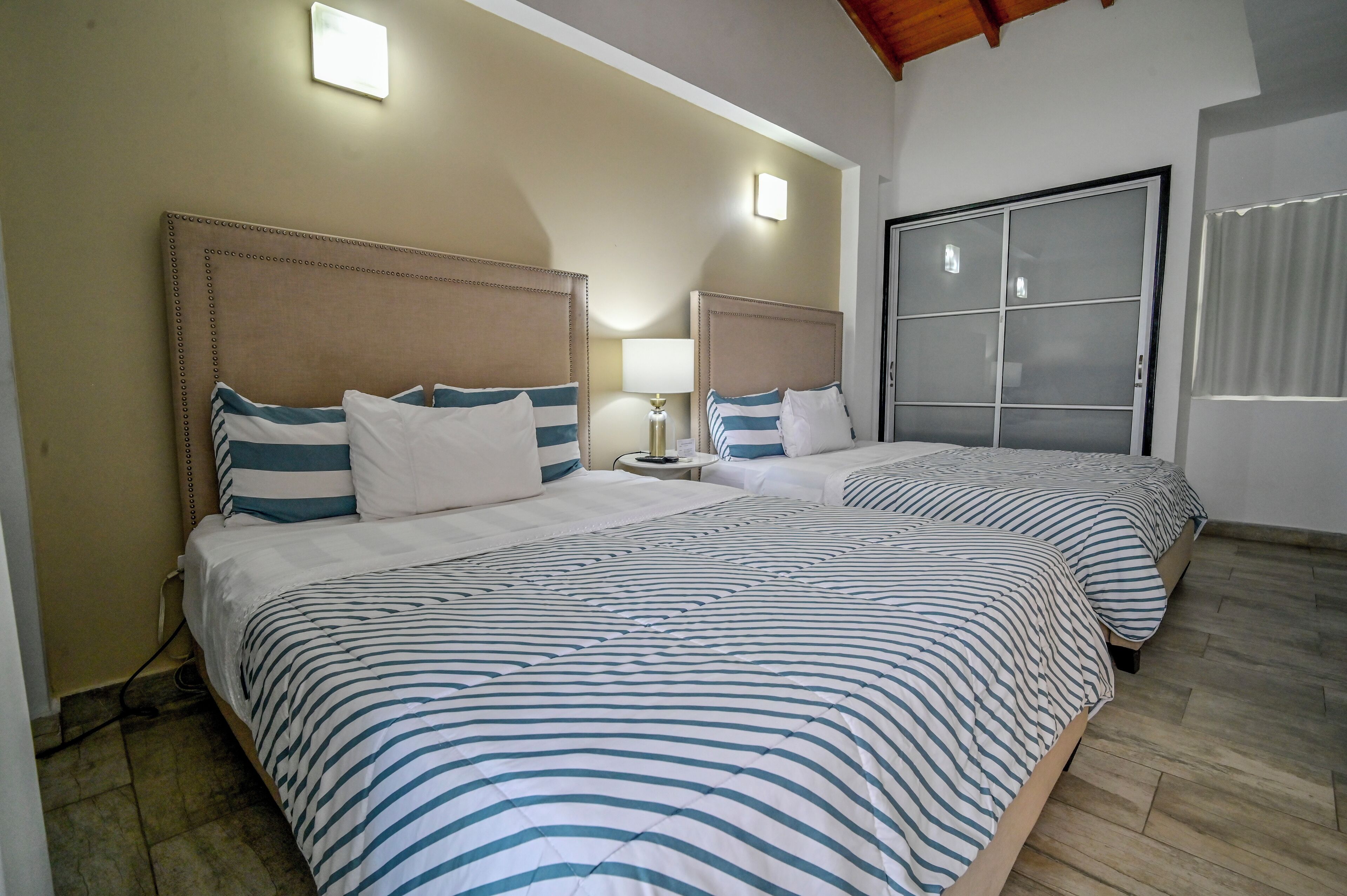 Quarto de hotel com duas camas, cada uma com roupa de cama listrada e cabeceiras, uma mesinha de cabeceira com abajur, e uma janela grande com vista.