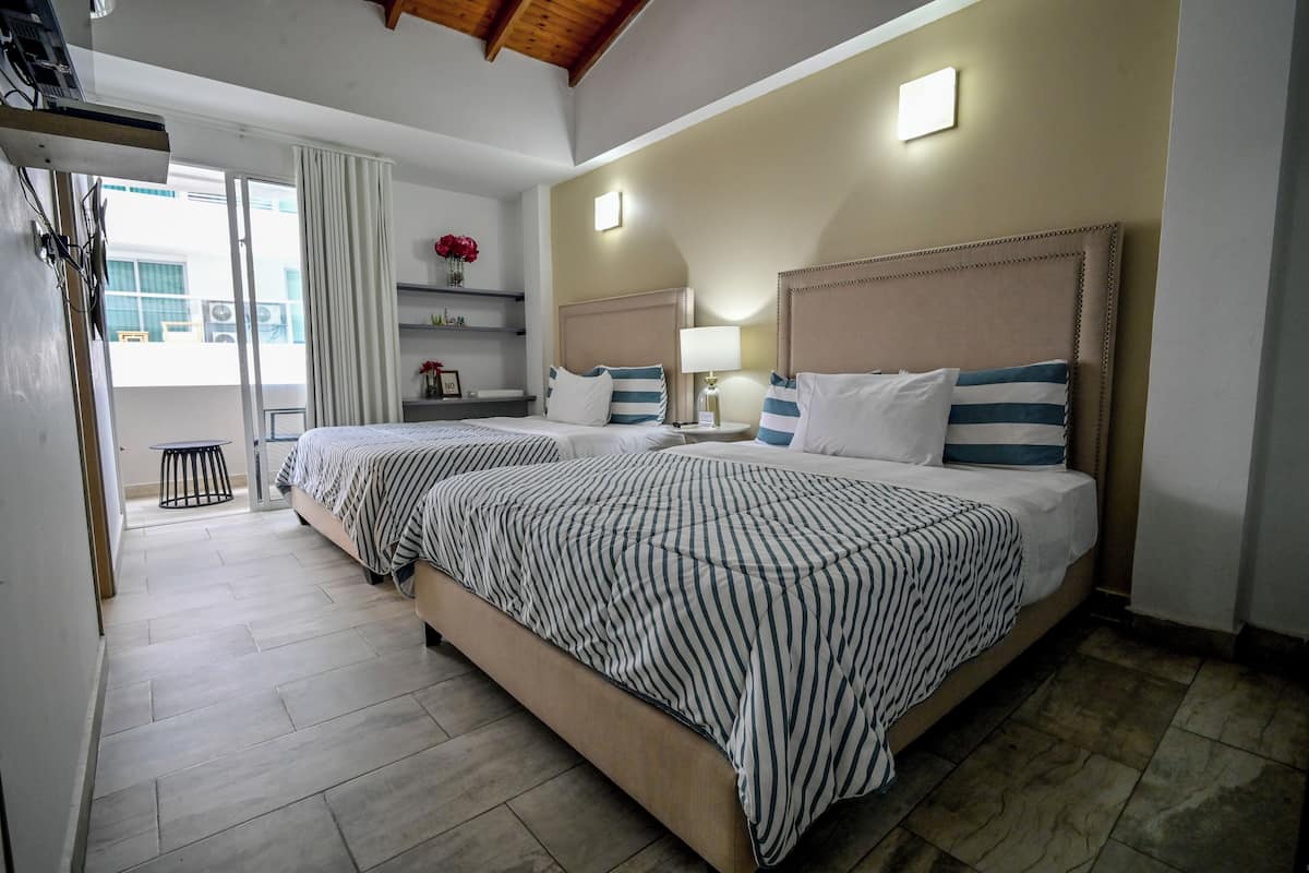 A hotel room with two beds, a balcony, a small table, and a wall-mounted shelf with flowers.