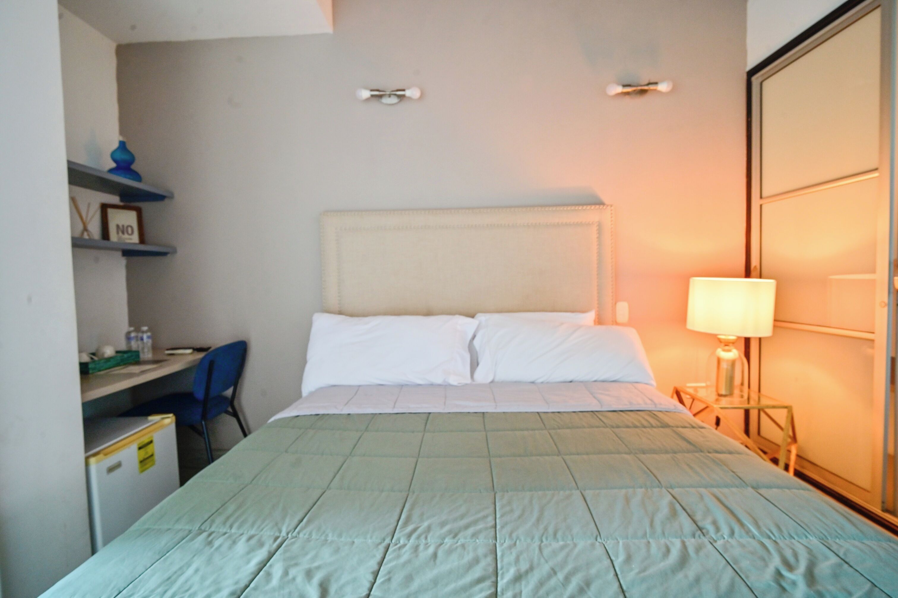 Quarto de hotel com cama, escrivaninha, cadeira, luminária e guarda-roupa.