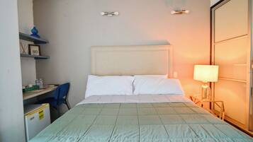 Quarto de hotel com cama, escrivaninha, cadeira, luminária e guarda-roupa.
