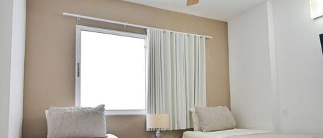 A hotel room with two beds, a ceiling fan, a window with curtains, and a lamp.