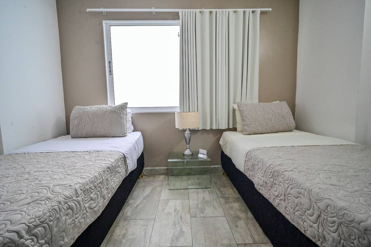 A hotel room with two beds, a window with curtains, a bedside table with a lamp, and a small sign.