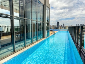 A rooftop swimming pool with glass walls, a view of the city skyline, and a wooden deck area.