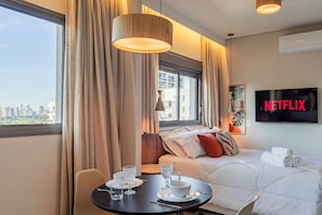 A hotel room with a bed, a TV displaying 'NETFLIX,' a dining table with glasses and a bowl, and a view of the city through the window.