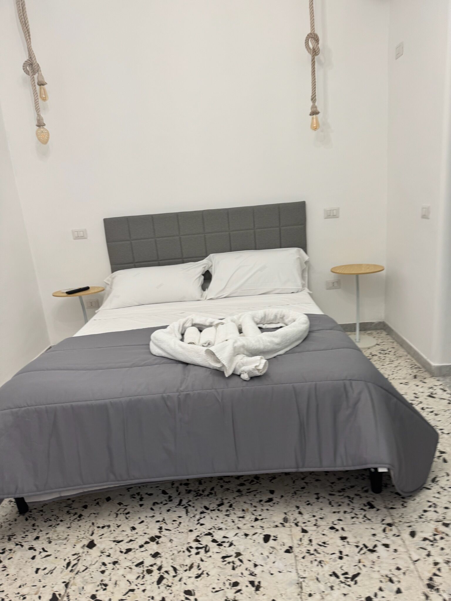 Una cama bien hecha con colcha gris y almohadas blancas, una mesita de noche con un control remoto y una mesa redonda con tapa trenzada.