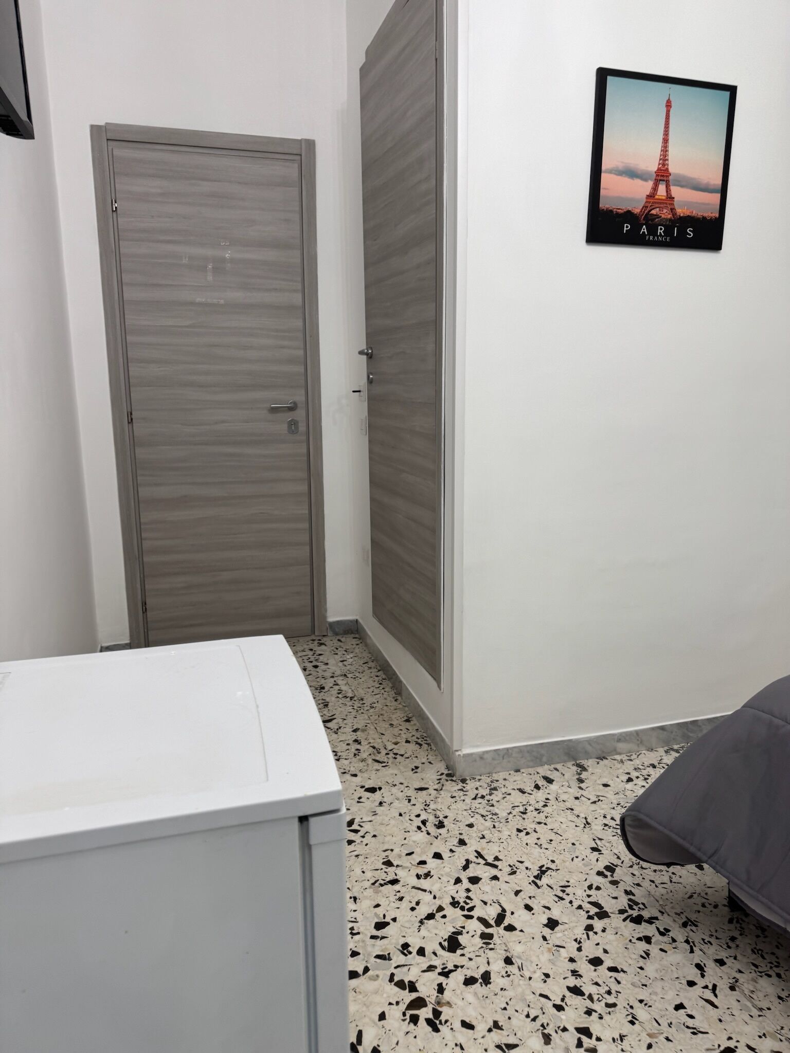 A room with a white washing machine, a grey wardrobe, and a bed with a grey blanket. A poster of the Eiffel Tower is on the wall.