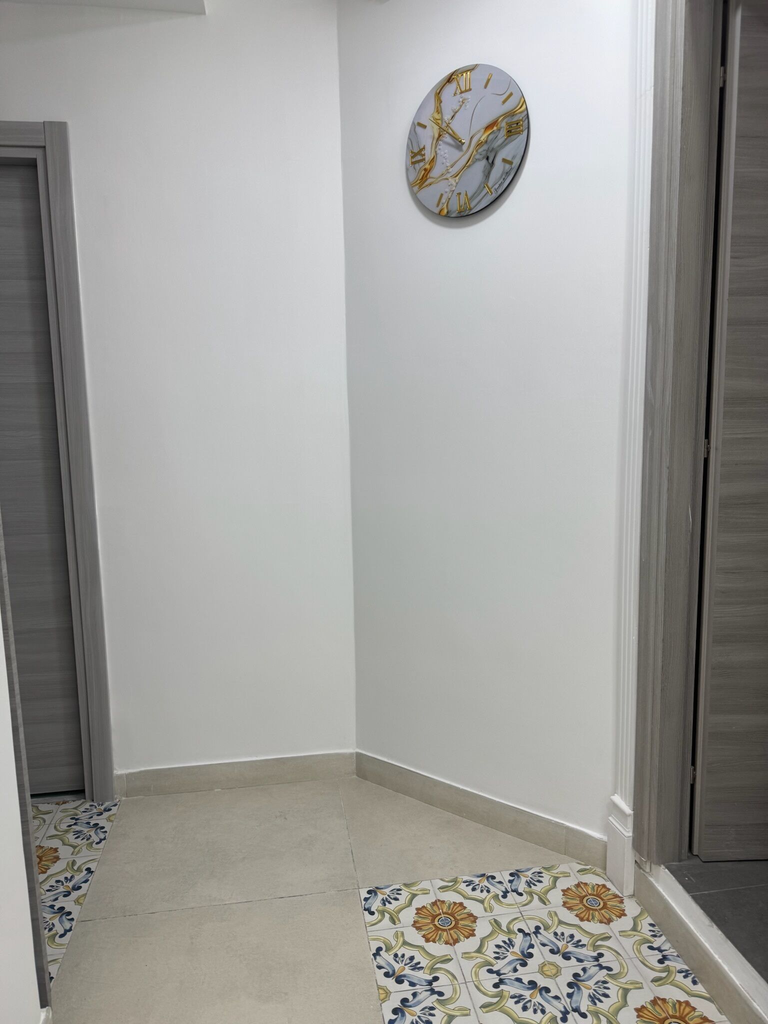 A hallway with a tiled floor, a plain white wall, and a clock with Roman numerals.