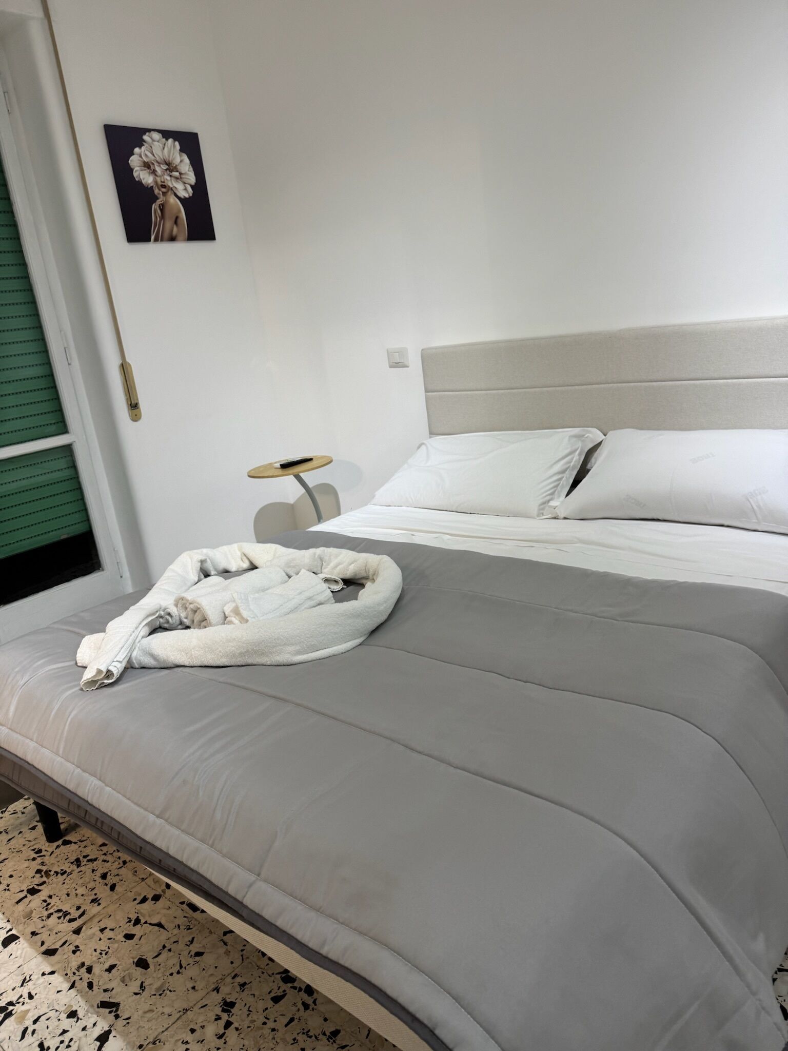 A neatly made bed with a grey duvet, a white pillow, and a white bathrobe on the bed. There is a small round table with a lamp beside the bed. A painting is visible on the wall above the bed.