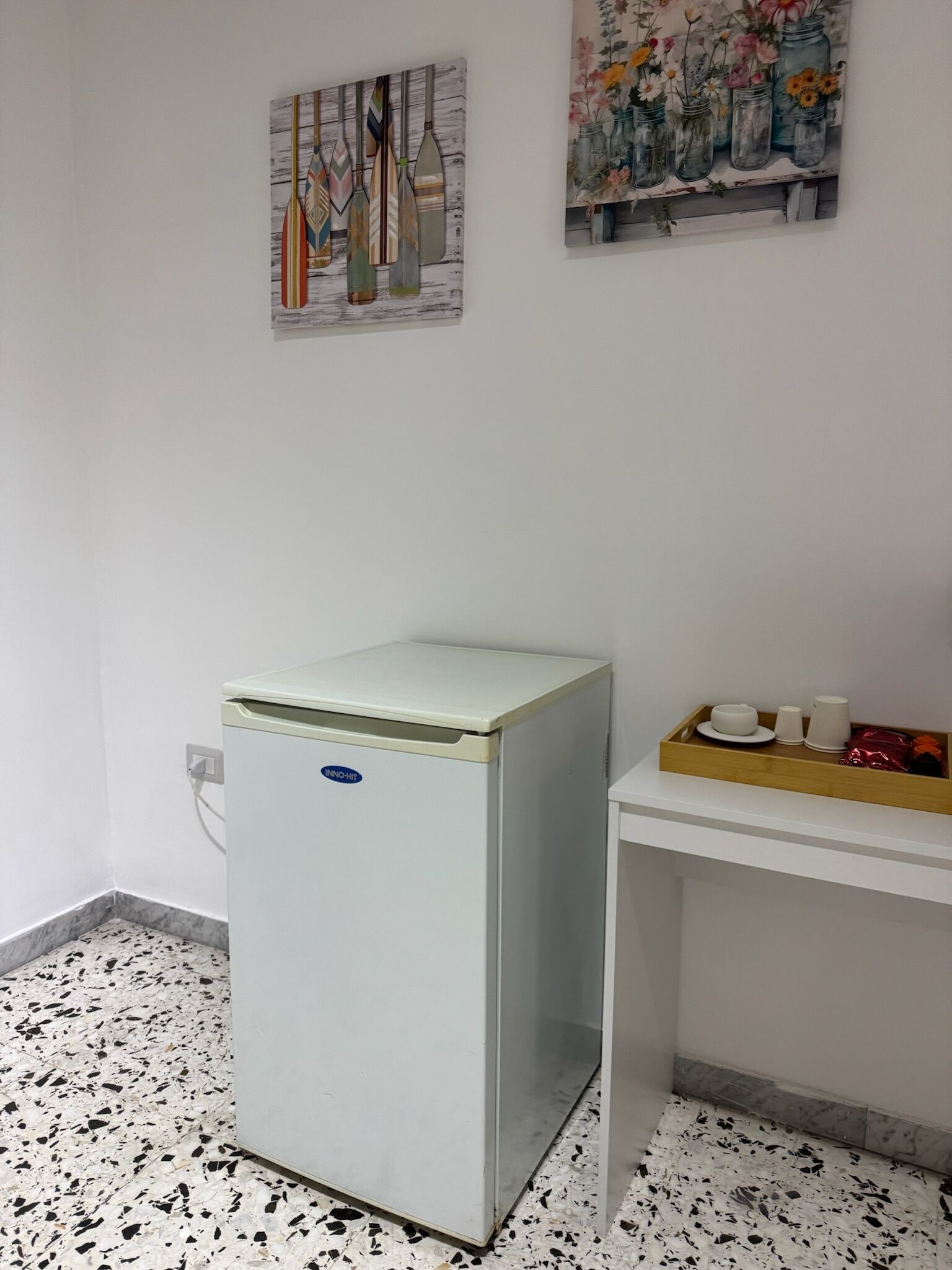 A small room with a white refrigerator, a white desk with a tray of cups and a small plant, and two wall-mounted artworks depicting bottles and flowers.