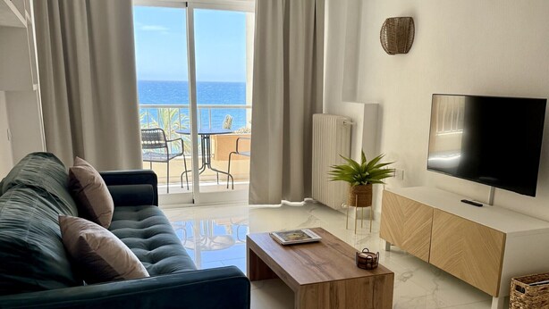 Un salon moderne avec un canapé, une table basse, un téléviseur et une vue sur la mer grâce à de grandes fenêtres.