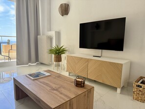Ein modernes Wohnzimmer mit Flachbildfernseher, Holzmöbeln, einer Topfpflanze und Blick ins Freie.