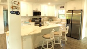 A modern kitchen with white cabinets, a stainless steel refrigerator, a marble countertop, and a bar area with two stools. - Lakefront Oasis w/ Heated Pool 2-Minutes Walk to Beach! (Holmes Beach)