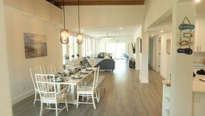 A spacious dining area with a long table, white chairs, and pendant lighting. A kitchen is visible in the background with a modern design. - Lakefront Oasis w/ Heated Pool 2-Minutes Walk to Beach! (Holmes Beach)