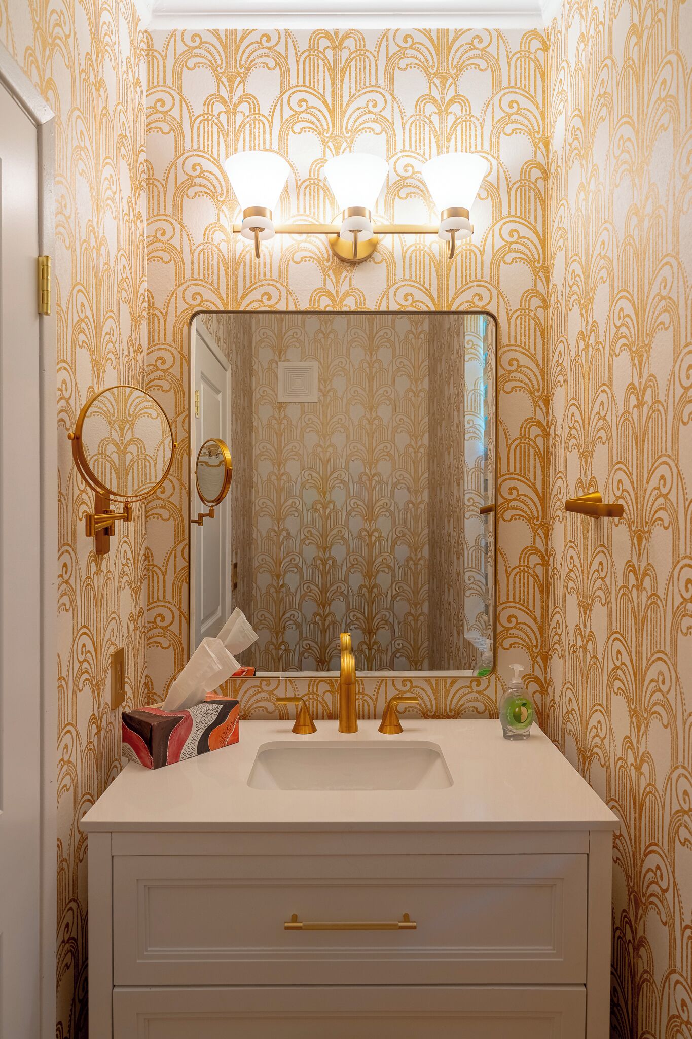 A bathroom with a white vanity, gold fixtures, and patterned wallpaper.
