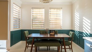 A dining area with a wooden table and four chairs, two windows with blinds, and a chandelier. - Beautiful 2-bedroom condo in cool San Diego close to the mall! (San Diego)
