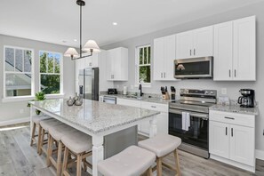 A modern kitchen with white cabinets, a large island with stools, stainless steel appliances, and a large window providing natural light. - Southern Comfort `B` | Near USC Downtown (Columbia)