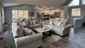 Ein großzügiges Wohnzimmer mit einer großen, grauen Eckcouch, einem Couchtisch und einem Essbereich mit Tisch und Stühlen. Das Zimmer verfügt über hohe Decken und große Fenster, die natürliches Licht hereinlassen.