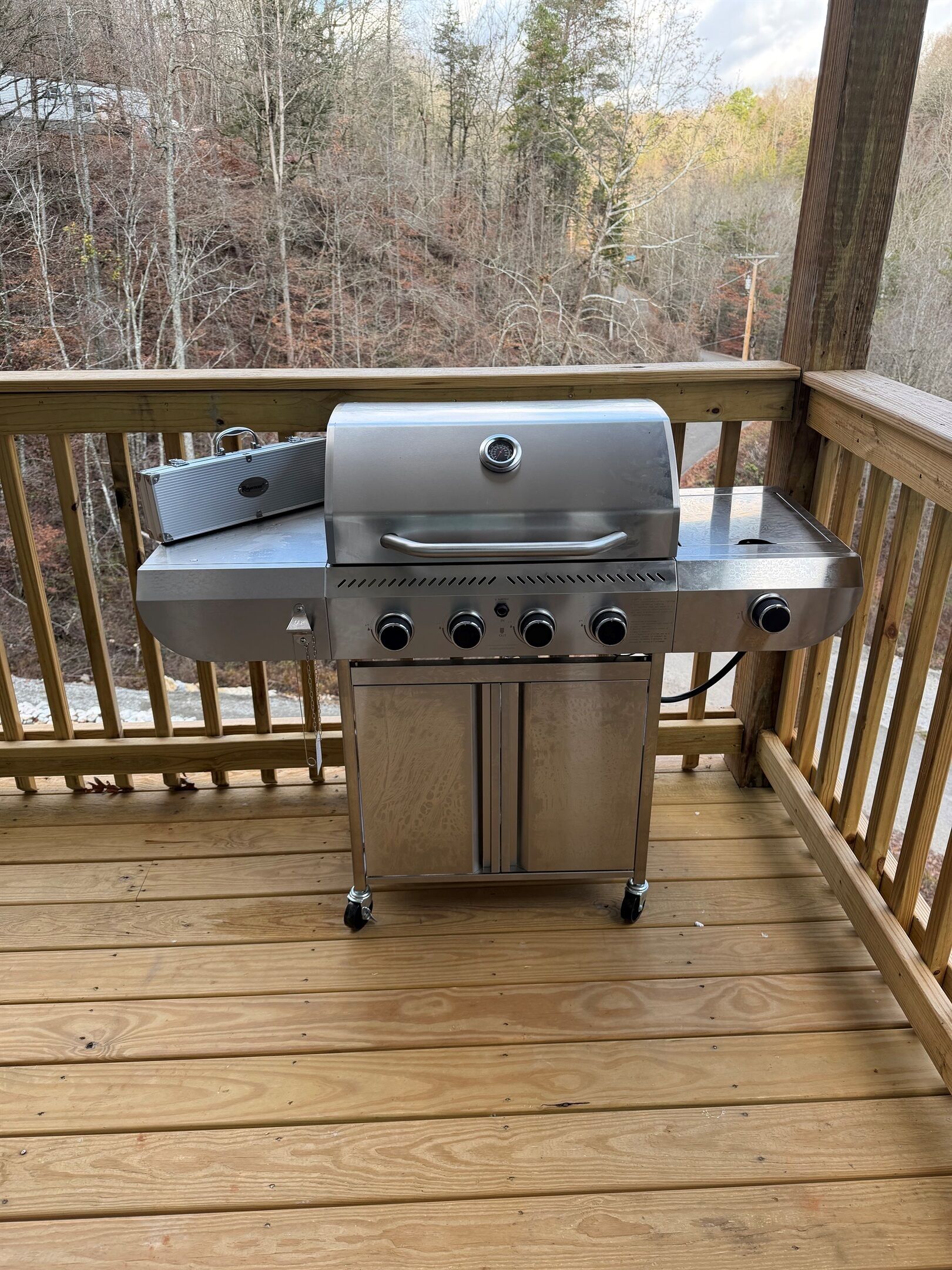 Ein Grill aus Edelstahl auf einer Holzterrasse mit Geländer, vor einer Baumkulisse.