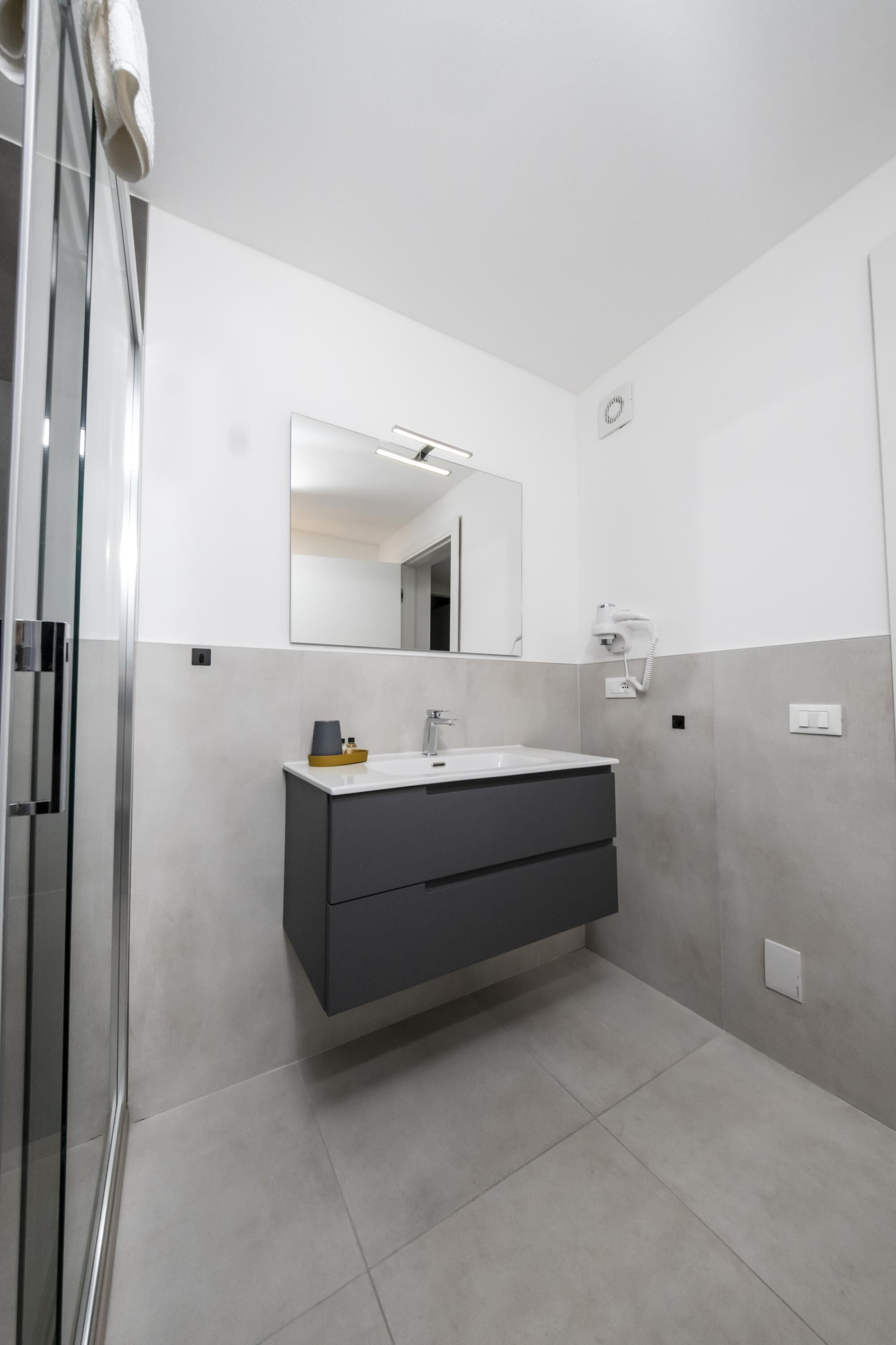 Une salle de bain moderne avec une douche à l’italienne en verre, un lavabo blanc avec un meuble sombre en dessous, et un grand miroir au-dessus du lavabo.