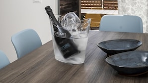 Une pièce avec une table en bois, deux chaises, une bouteille de champagne et un verre. Une fenêtre offre une vue sur un bâtiment et des collines.