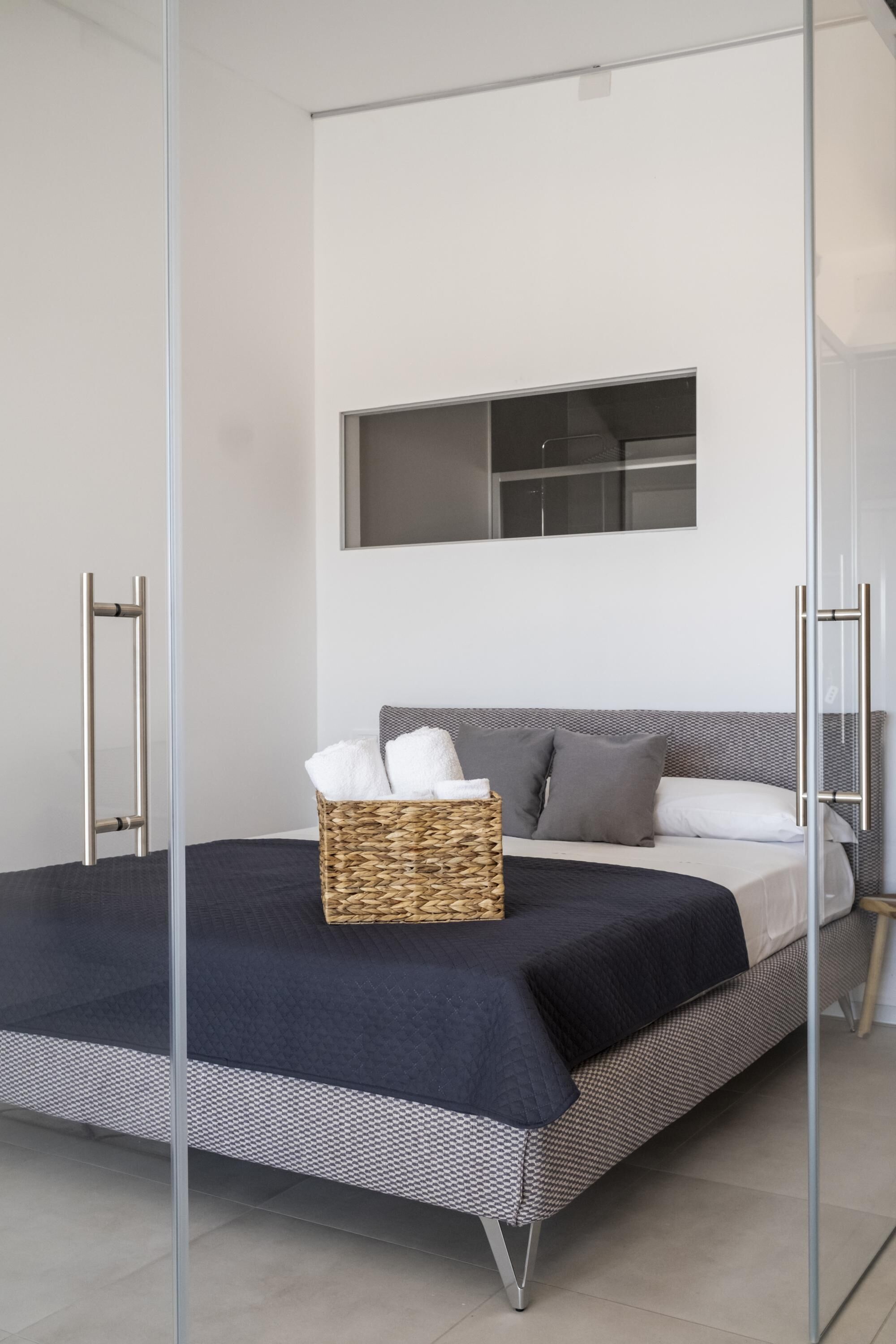 A modern bedroom with a large bed, a bedside table, a basket of towels, and a window above the bed.