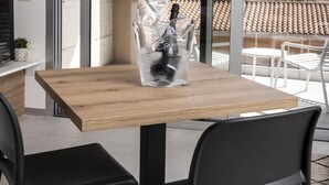 Un espace repas moderne avec une table en bois et des chaises noires, un vase en verre contenant une bouteille, et une vue sur un toit recouvert de tuiles et des montagnes, visible à travers de grandes portes vitrées.