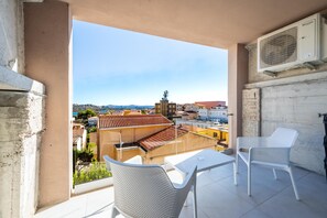 Ein Balkon mit zwei weißen Stühlen und einem Tisch, mit Blick auf eine Stadtlandschaft mit Gebäuden und blauem Himmel.