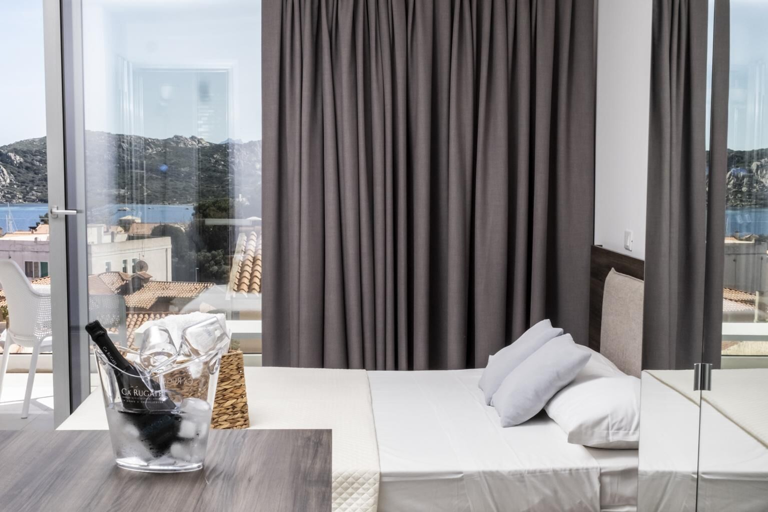 A hotel room with a bed, a glass table with a bottle, and a view of buildings and hills.