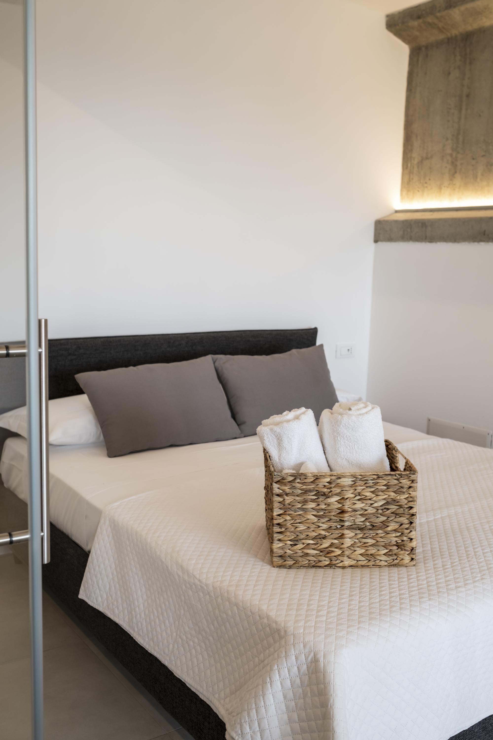 A neatly made bed with a white quilt, two grey pillows, and a woven basket containing two white towels.