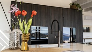 A modern kitchen with a view of the sea, a wooden cutting board, a gold vase with red flowers, and a decorative metal container. - Attico Salia - La Maddalena (La maddalena)