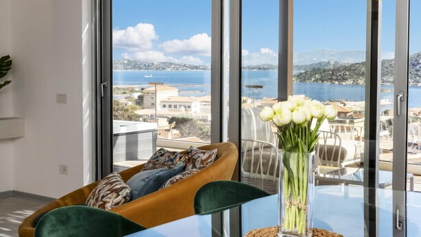 A modern dining area with a glass table, green upholstered chairs, and a view of the sea through large windows. - Attico Salia - La Maddalena (La maddalena)