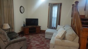 Una sala de estar con un televisor sobre un mueble de madera, un sofá, una alfombra estampada y una escalera.