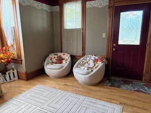 Una habitación con dos sillones blancos, suelo de madera, alfombra estampada, puerta decorativa y ventana con cortinas de encaje.