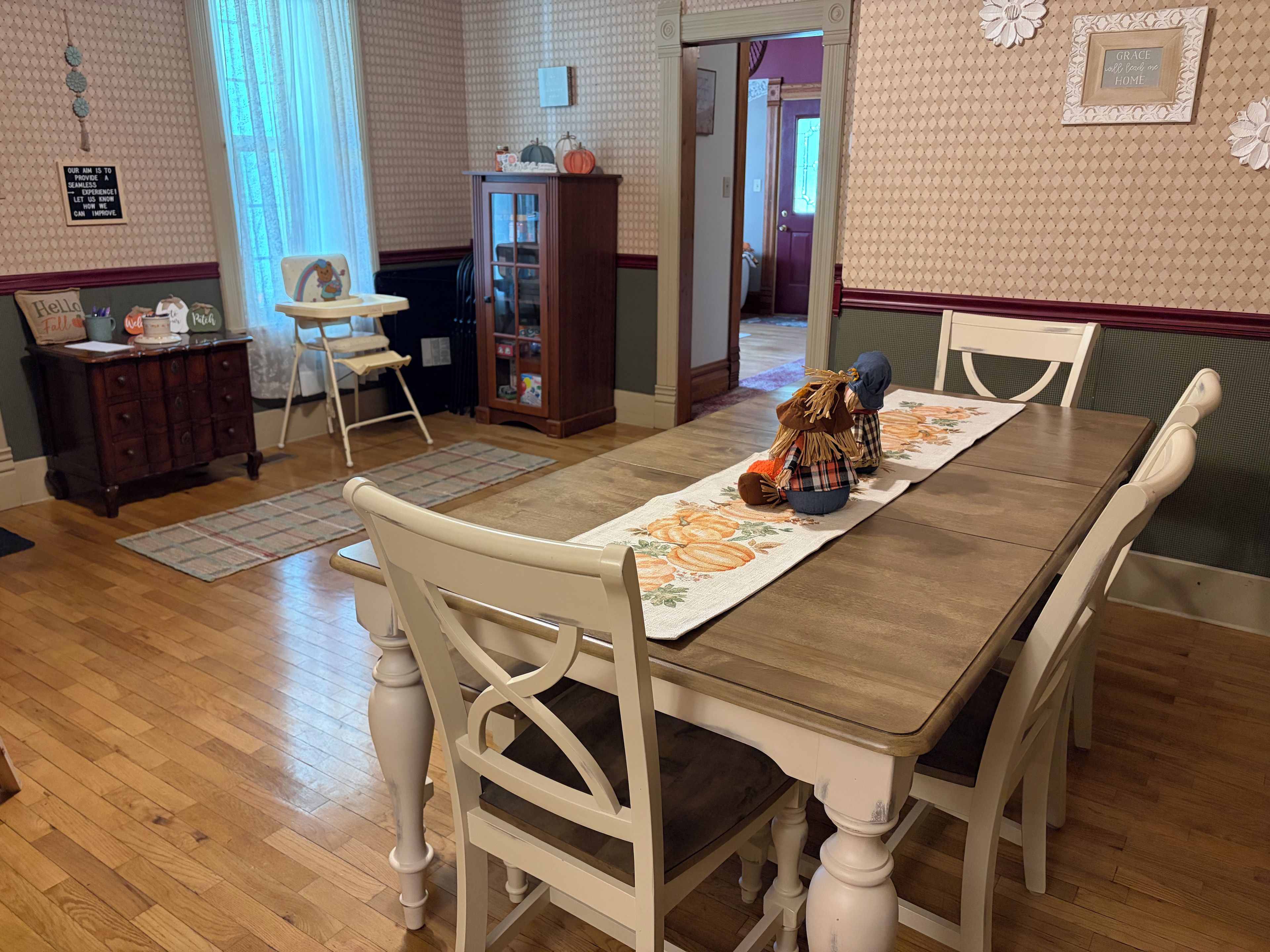 A dining room with a wooden table, white chairs, a floral table runner, and a cabinet with decorative items.