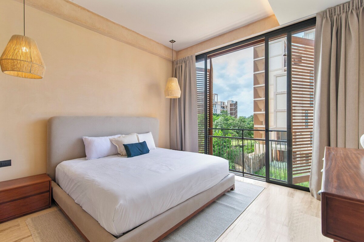Ein modernes Schlafzimmer mit einem großen Bett, einem Holznachttisch, einer Pendelleuchte und einem Balkon mit Blick auf Gebäude und Grünflächen.